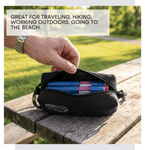 Hand opening a black zippered pouch containing blue and red items on a wooden surface with text about its uses. 