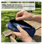 A blue zippered pouch with the brand name 'BREEZY PACKS' printed on the front, being opened by two hands outdoors on a wooden picnic table with a grassy park background. A French text overlay is visible at the top. 